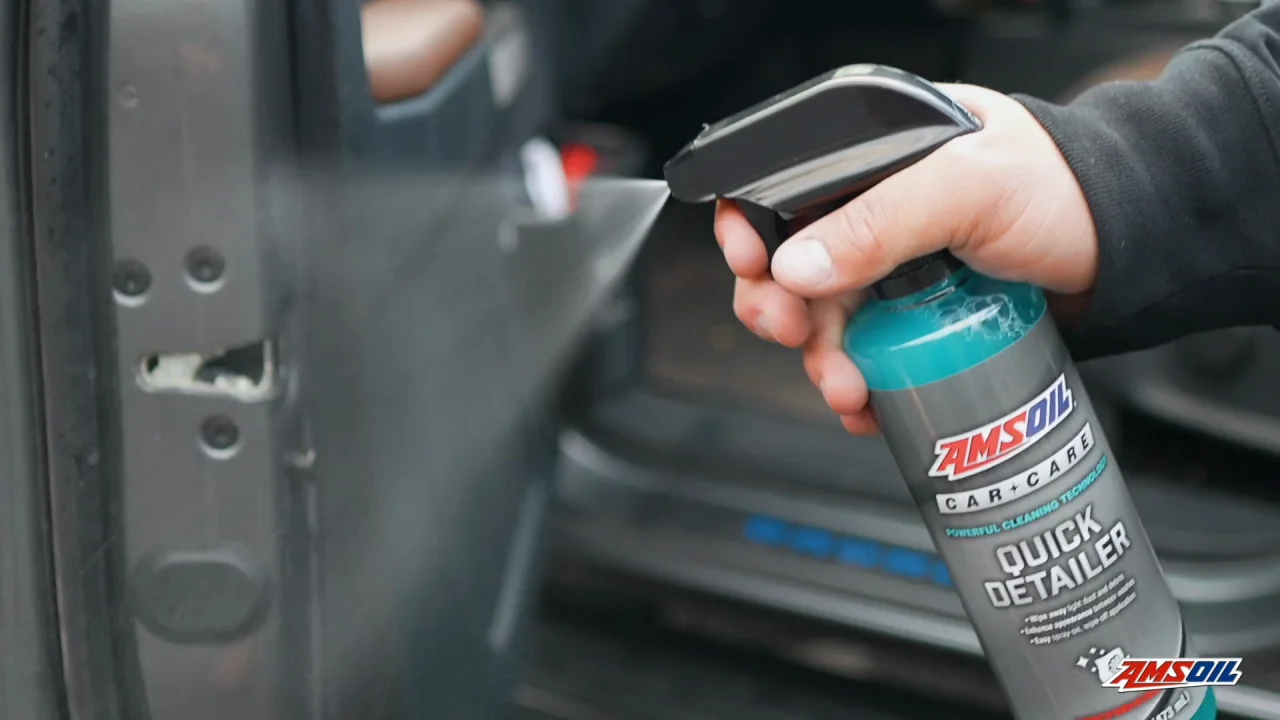 A hand holding and spraying a bottle of AMSOIL quick detailer into a dirty door jam of a front drivers side door of a vehicle.