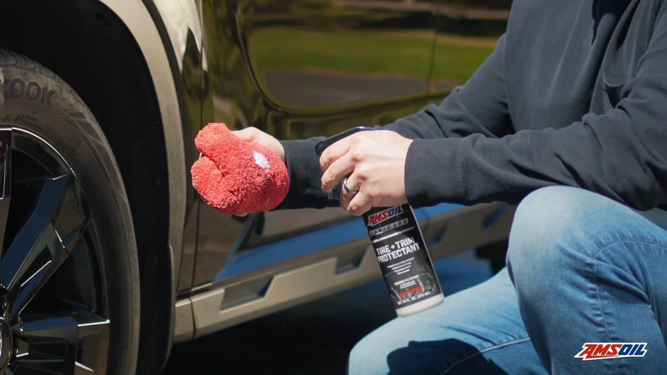 Hands holding and spraying a bottle of AMSOIL Tire and Trim Protectant into a red microfiber towel