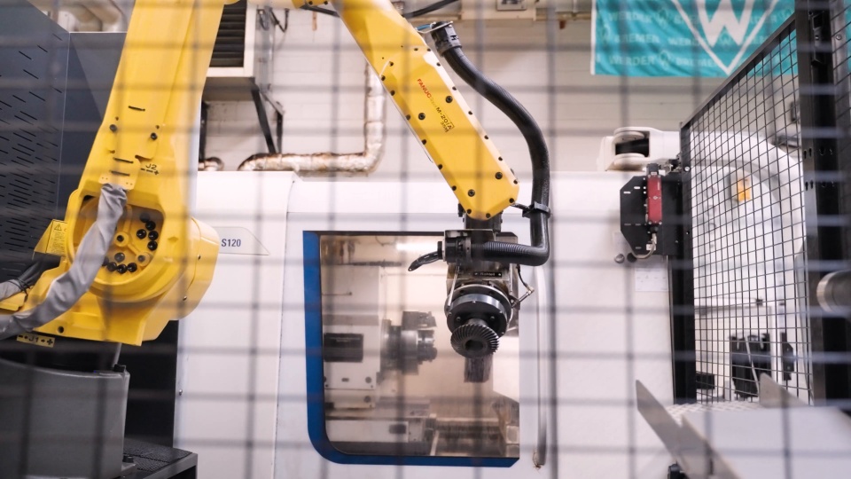 Still from the Tandler recruiting clip: a yellow robot arm at a machining center
