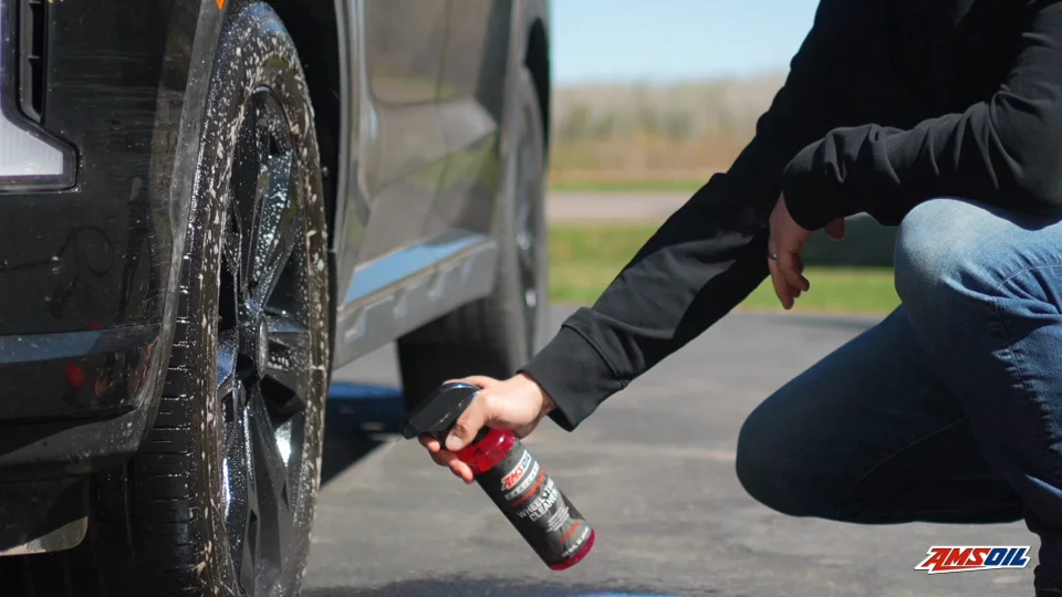 A hand holding and spraying a bottle of AMSOIL wheel and tire cleaner on to a black wheel of a car.  The contents of the bottle are red.